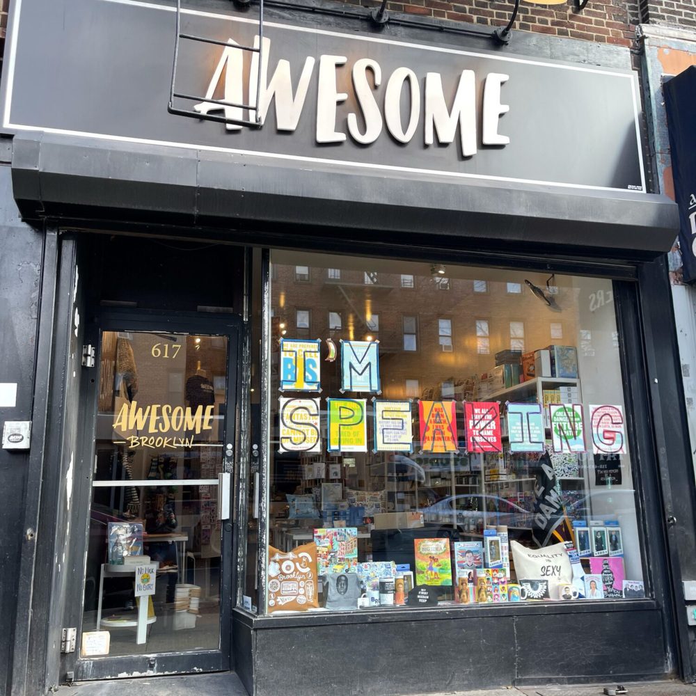 Storefront of "Awesome Brooklyn" with books and games displayed in the window, decorated with colorful "I'M SPEAKING" letters.