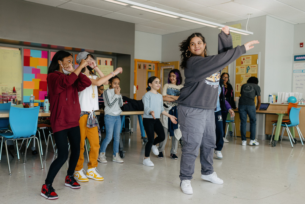 A group of children dancing enthusiastically in a classroom with colorful chairs, displaying various playful poses and expressions.