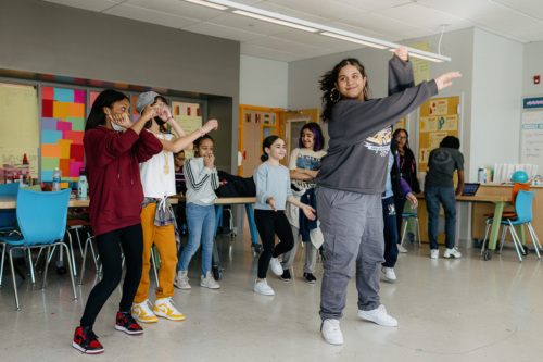 A group of children dancing enthusiastically in a classroom with colorful chairs, displaying various playful poses and expressions.