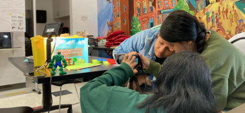 Two people focus on operating a camera on a tripod indoors, near a table with small models and a colorful backdrop.