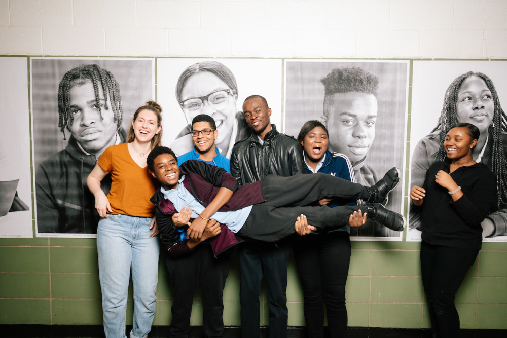 A group of seven people smiling and posing in front of black and white portraits. One person is being held horizontally by others. The background has a tiled wall.