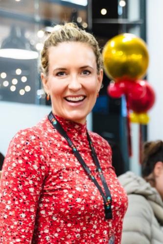 Person wearing a red patterned top and lanyard stands indoors, smiling. Background features balloons and blurred lights.