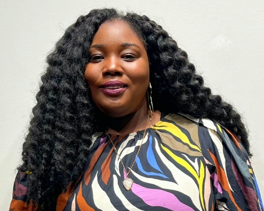 Woman with long curly hair wearing a colorful striped dress and a pendant necklace, standing against a plain background.