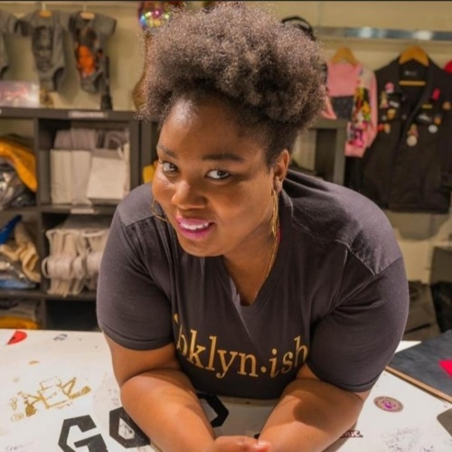 Person with a natural hairstyle and gold hoop earrings leans on a table, wearing a black "Bklyn.ish" shirt. Shelves with various items are in the background.