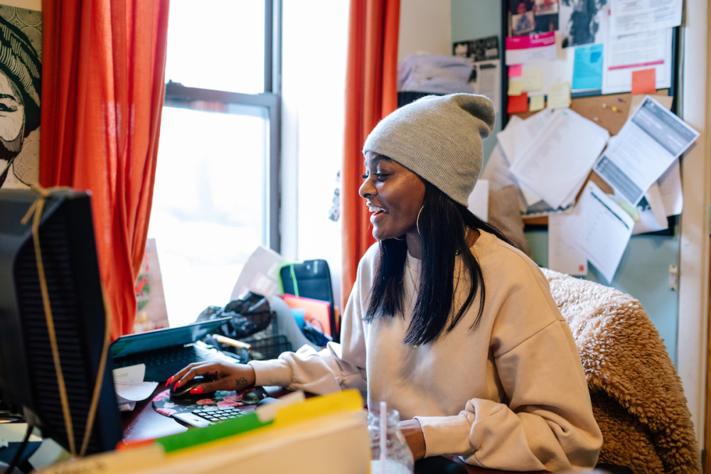 Person wearing a beanie and sweater works at a computer in a cozy, cluttered room with orange curtains and a bulletin board.