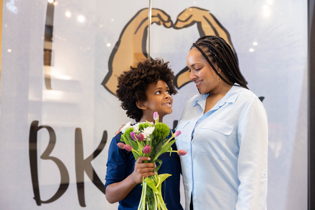 A child holding a bouquet of flowers stands next to an adult, both smiling, in front of a mural showing hands forming a heart shape.