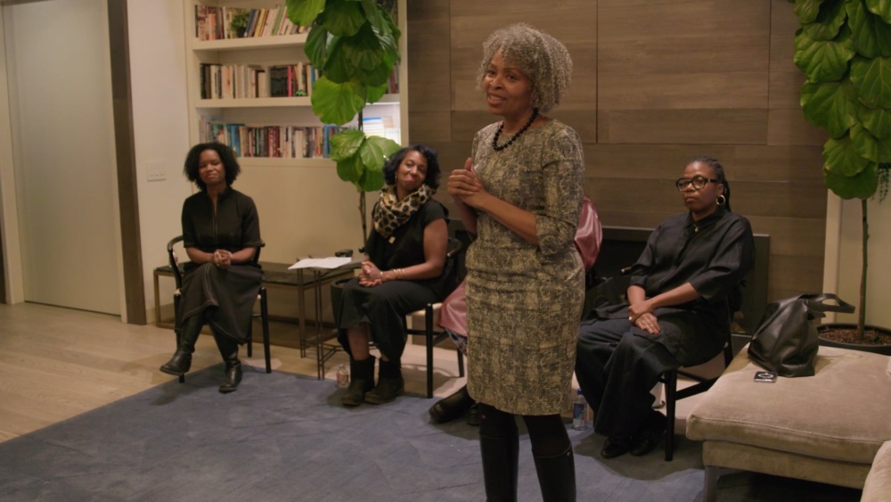 A woman speaks to a small seated audience in a room with bookshelves and large plants.