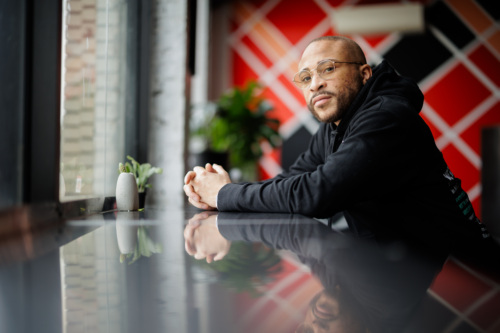 A person wearing glasses and a black hoodie leans on a reflective table by a window, with plants and a geometric red and black wall in the background.