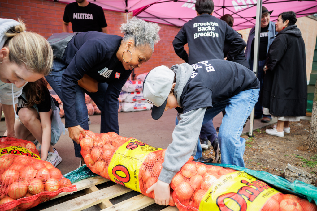 Two people lift a large mesh bag of onions onto a wooden pallet at an outdoor food distribution event, while others work in the background under a pink tent.