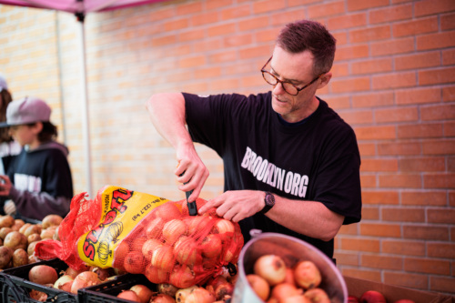 A man wearing glasses and a "BROOKLYNORG" shirt opens a large red mesh bag of onions at an outdoor market stall with more onions on the table.