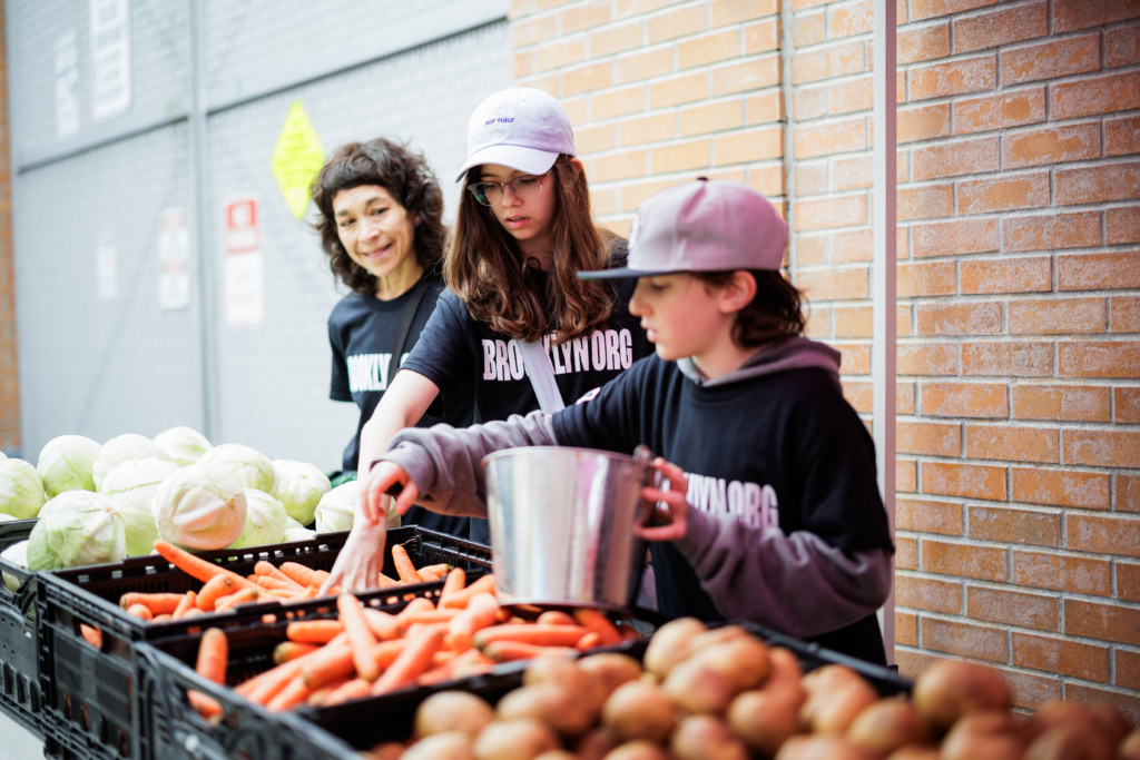 Three people wearing matching shirts and hats sort carrots, onions, and cabbage at an outdoor produce stand near a brick wall.