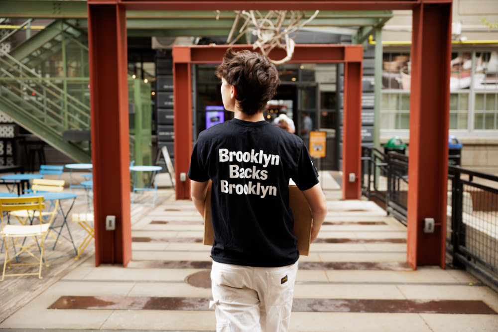 A person with short brown hair, wearing a "Brooklyn Backs Brooklyn" t-shirt and white pants, walks through an outdoor urban area carrying a box.