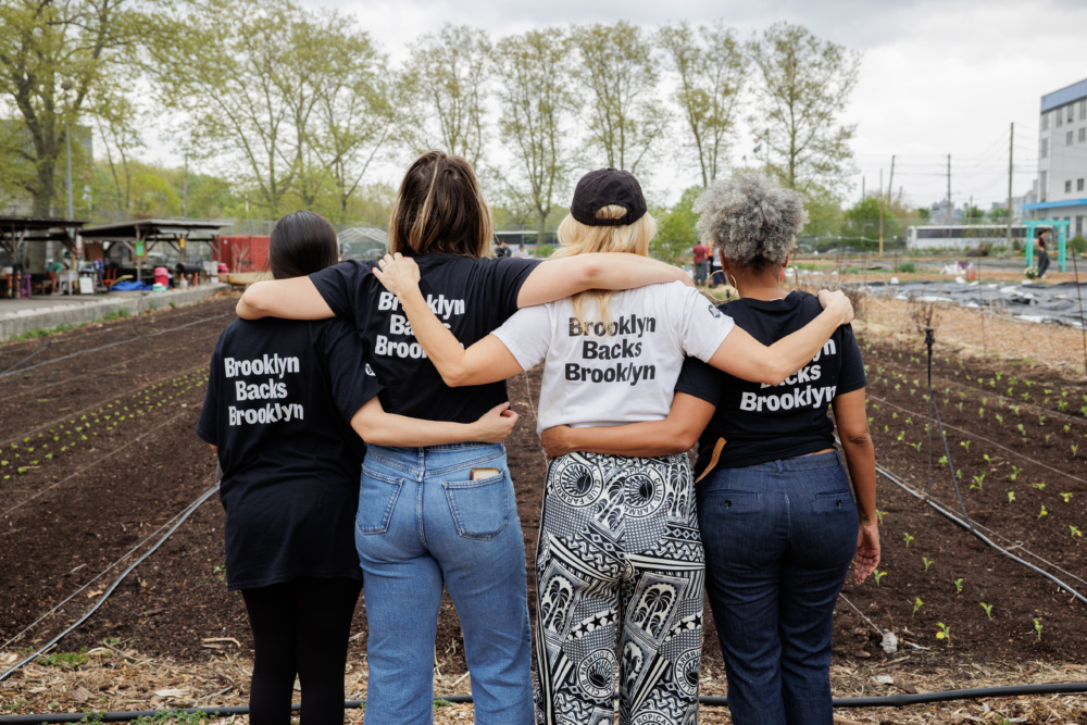 Four people standing arm-in-arm in front of crop rows, wearing "Brooklyn Backs Brooklyn" shirts, facing away from the camera on a cloudy day.