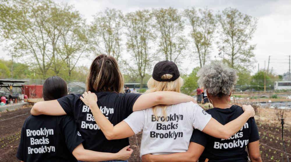 Four people standing arm-in-arm in front of crop rows, wearing "Brooklyn Backs Brooklyn" shirts, facing away from the camera on a cloudy day.