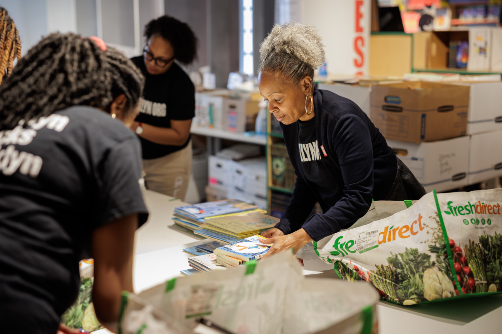 A woman organizes books on a table while others sort and pack items in a room filled with boxes and shopping bags.