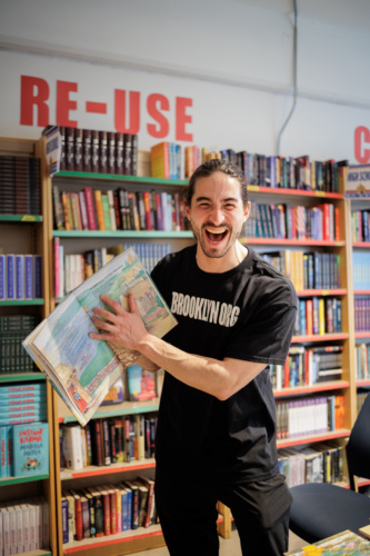 A man wearing a "BROOKLYN.ORG" t-shirt stands in a bookstore, smiling and holding an open illustrated book in front of bookshelves.