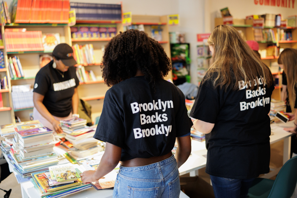 Several people wearing "Brooklyn Backs Brooklyn" shirts sort and organize stacks of books on tables in a bookstore or library.