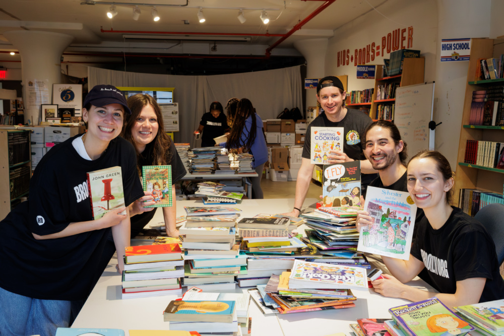 Five people smile while sorting and displaying children’s books at a table in a well-lit room lined with shelves filled with more books.
