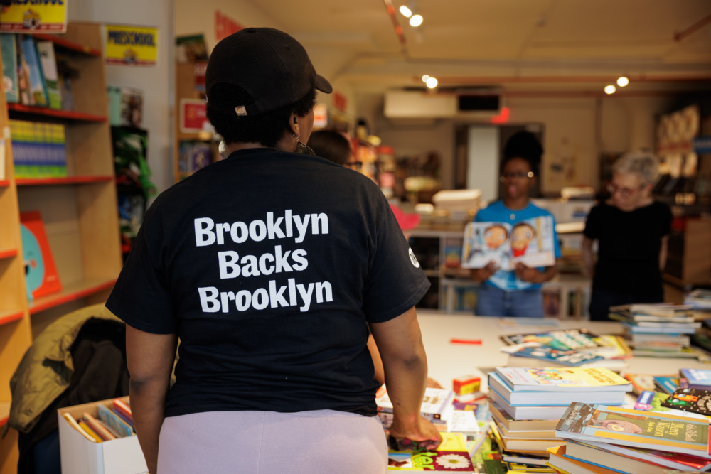 A person wearing a "Brooklyn Backs Brooklyn" shirt stands at a table covered with books in a bookstore, with others browsing in the background.