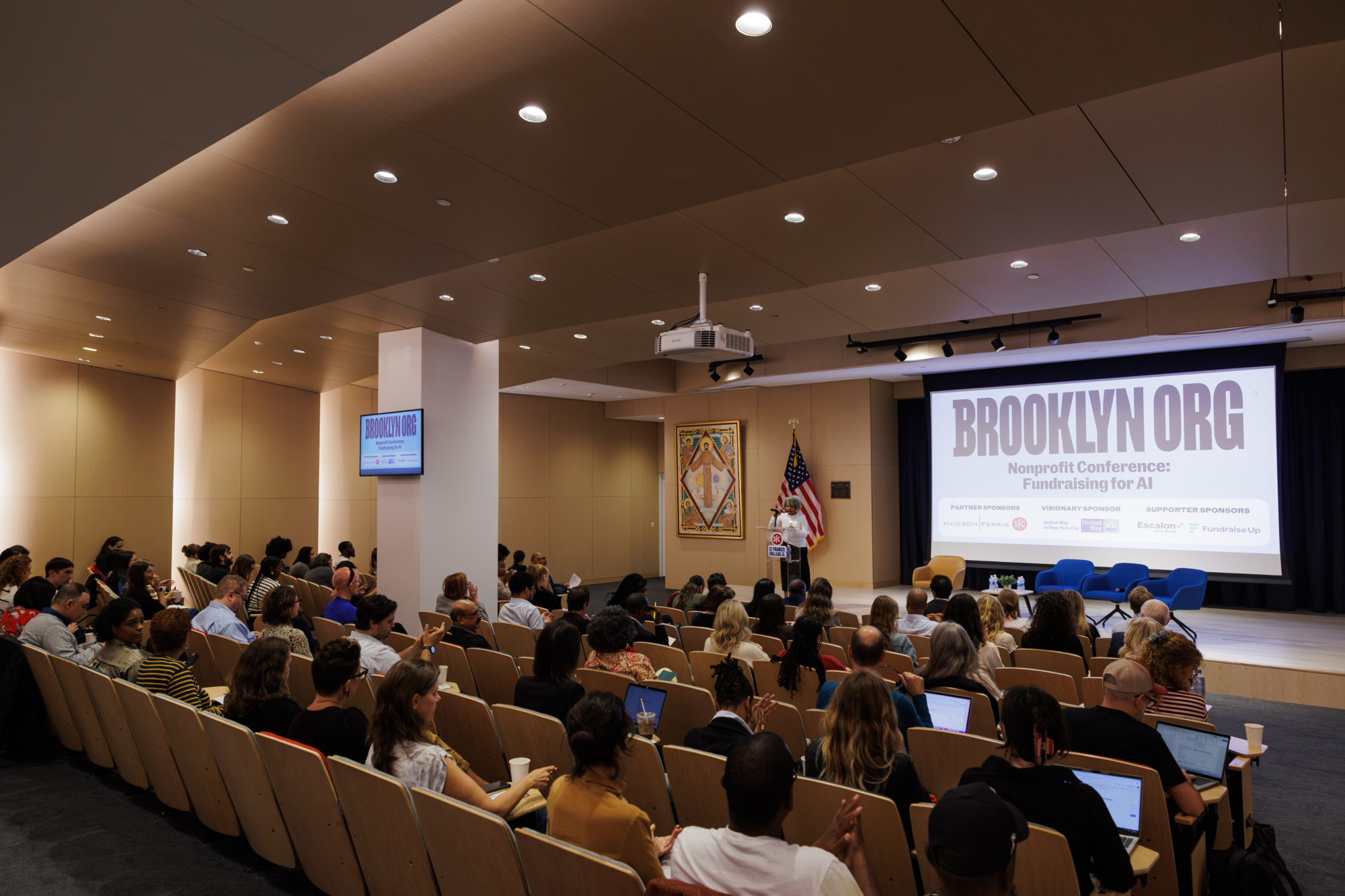 A speaker addresses an audience in an auditorium at the Brooklyn Org Nonprofit Conference, with event information displayed on a large screen behind them.