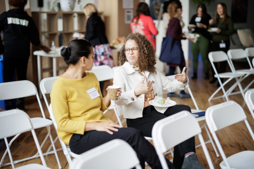 Two women seated on white chairs engage in conversation. One holds a coffee cup, the other gesticulates while holding a plate with food. Other people are visible in the background.
