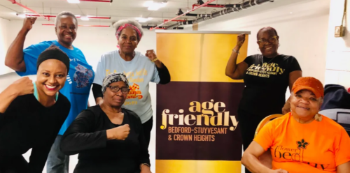 Six older adults pose with raised fists next to an "Age Friendly Bedford-Stuyvesant & Crown Heights" banner in a brightly lit indoor space.