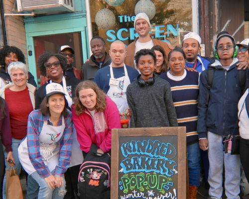 A group of people pose in front of a bakery pop-up. A sign reads "Kindred Bakery's Pop Up: Muffins, Cookies, Scones, Cakes, Breads." Balloons are visible in the background.