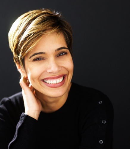 Woman with short, highlighted hair smiles broadly, wearing a black sweater and resting her hand on her neck against a dark background.