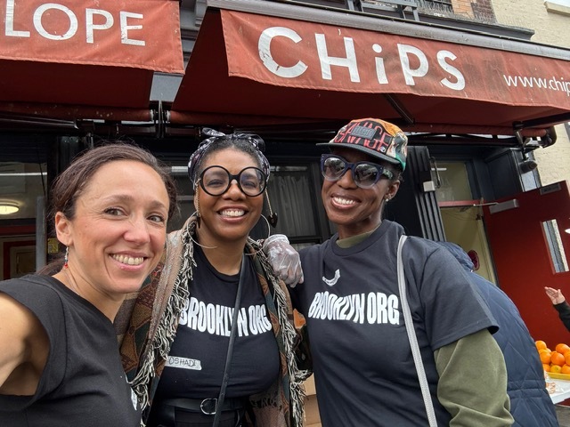 Three women wearing "BROOKLYN ORG" shirts smile for a photo outside a shop with a red "CHiPS" awning.