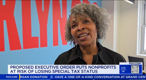 A woman with gray curly hair speaks in front of a blue wall with red text. A news ticker below discusses a proposed executive order affecting nonprofit tax status.