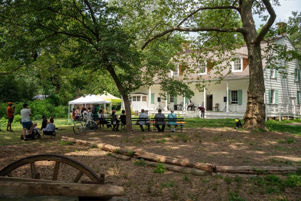 People gather on benches and lawn under trees near a large white house with a porch; a white tent is set up nearby on a sunny day.
