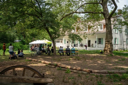 People gather on benches and lawn under trees near a large white house with a porch; a white tent is set up nearby on a sunny day.