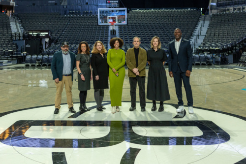 Seven individuals stand on a basketball court, arranged in a line for a group photo, facing the camera, with empty stadium seats visible in the background.