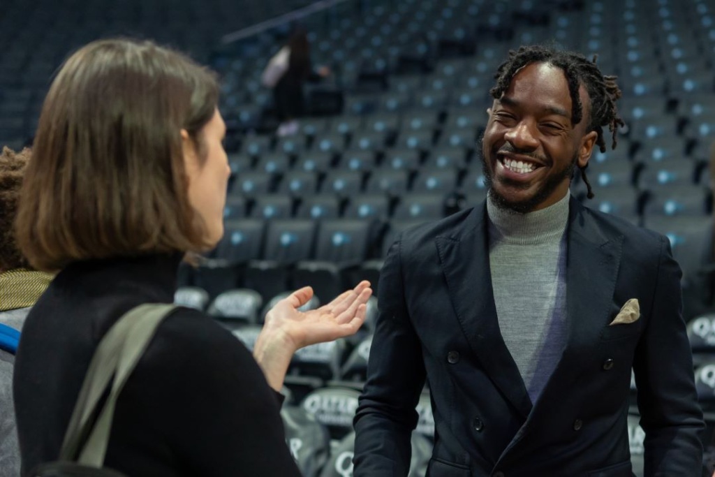Two people engaging in conversation in an empty stadium. The man is smiling, wearing a suit; the woman is gesturing with her hand. Rows of empty seats are visible in the background.
