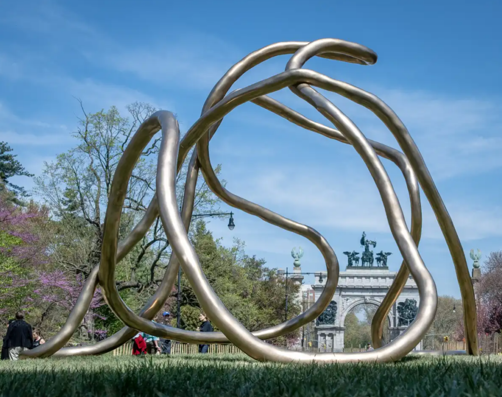 A large abstract bronze sculpture of looping, intertwined tubes sits on grass in a park, with trees and a stone monument visible in the background.