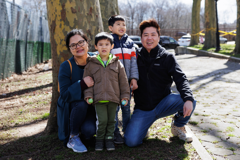 A group of four people, two adults and two children, pose and smile outdoors on a sunny day next to a tree in a park.