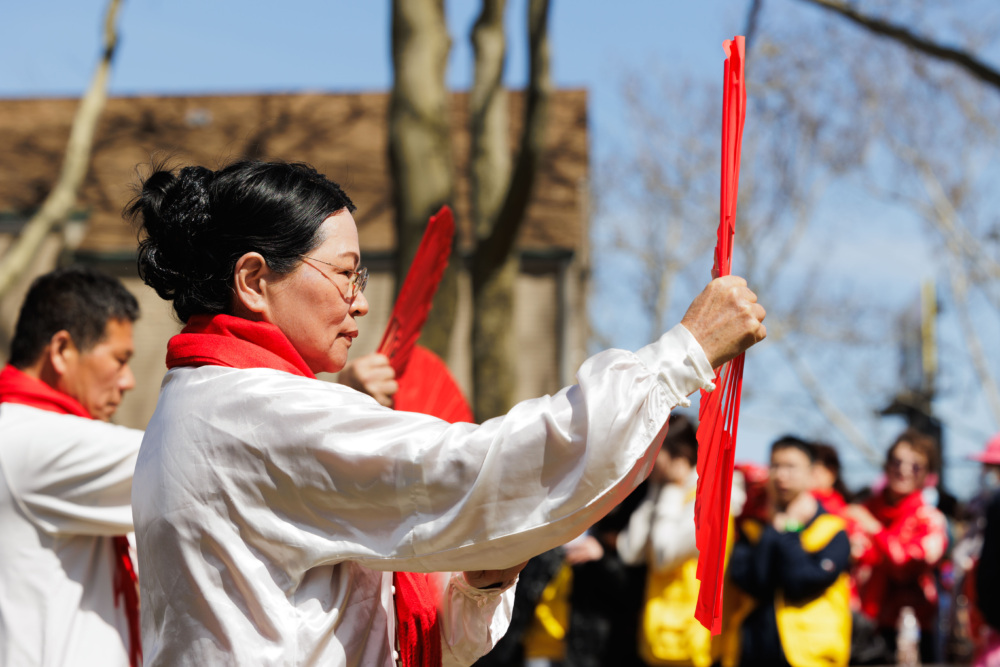 A woman in a white shirt and red scarf holds a red fan during an outdoor group event, with other participants visible in the background.