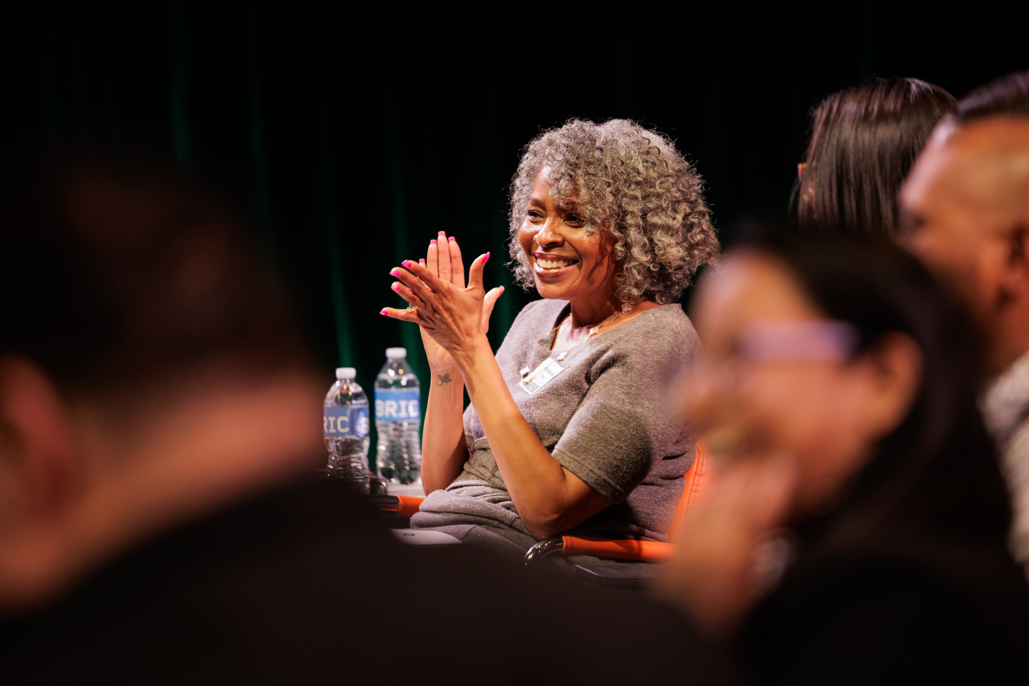 An older woman with curly gray hair smiles and claps while seated at an event, with water bottles on the table in front of her and other attendees visible in the foreground.