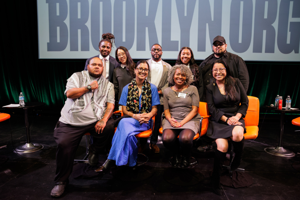 A group of ten people pose together on stage, seated and standing in front of an orange chair arrangement and a large screen displaying the text “BROOKLYN.ORG.”.