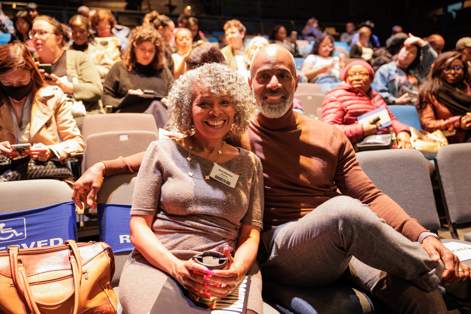 A smiling couple sits together in a crowded auditorium with people around them, some reading programs and wearing nametags.