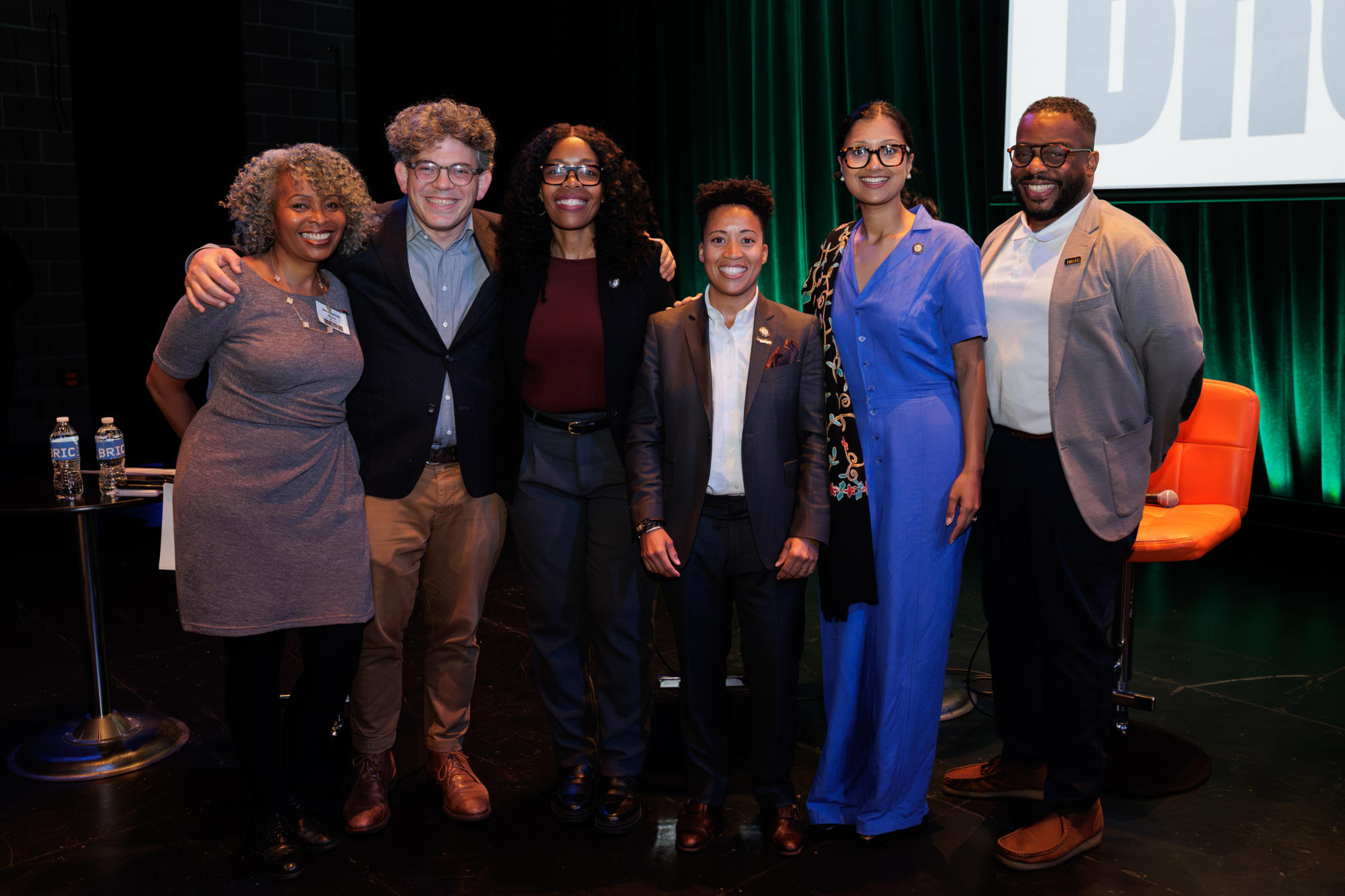 Six people stand and pose for a group photo on a stage with dark walls and green lighting, some smiling, dressed in business and business casual attire.