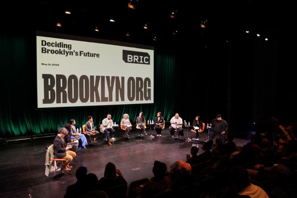 A panel of ten people sits on stage under a large screen reading "Deciding Brooklyn's Future BRIC BROOKLYN.ORG," addressing an audience in a dark theater.