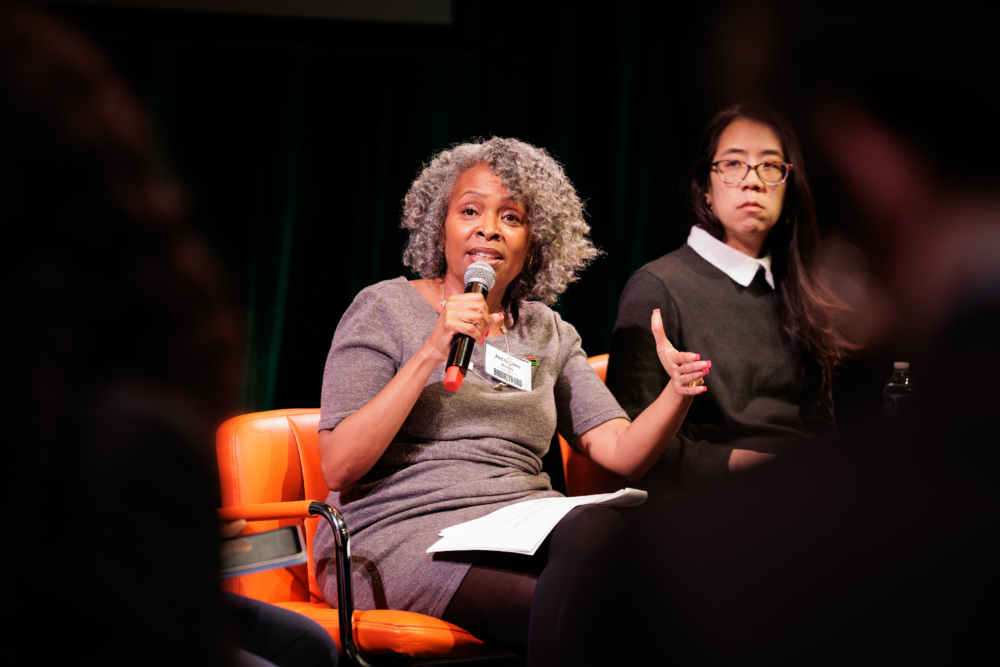 A woman with gray curly hair speaks into a microphone while seated on a panel, with another woman sitting beside her, both facing an audience.