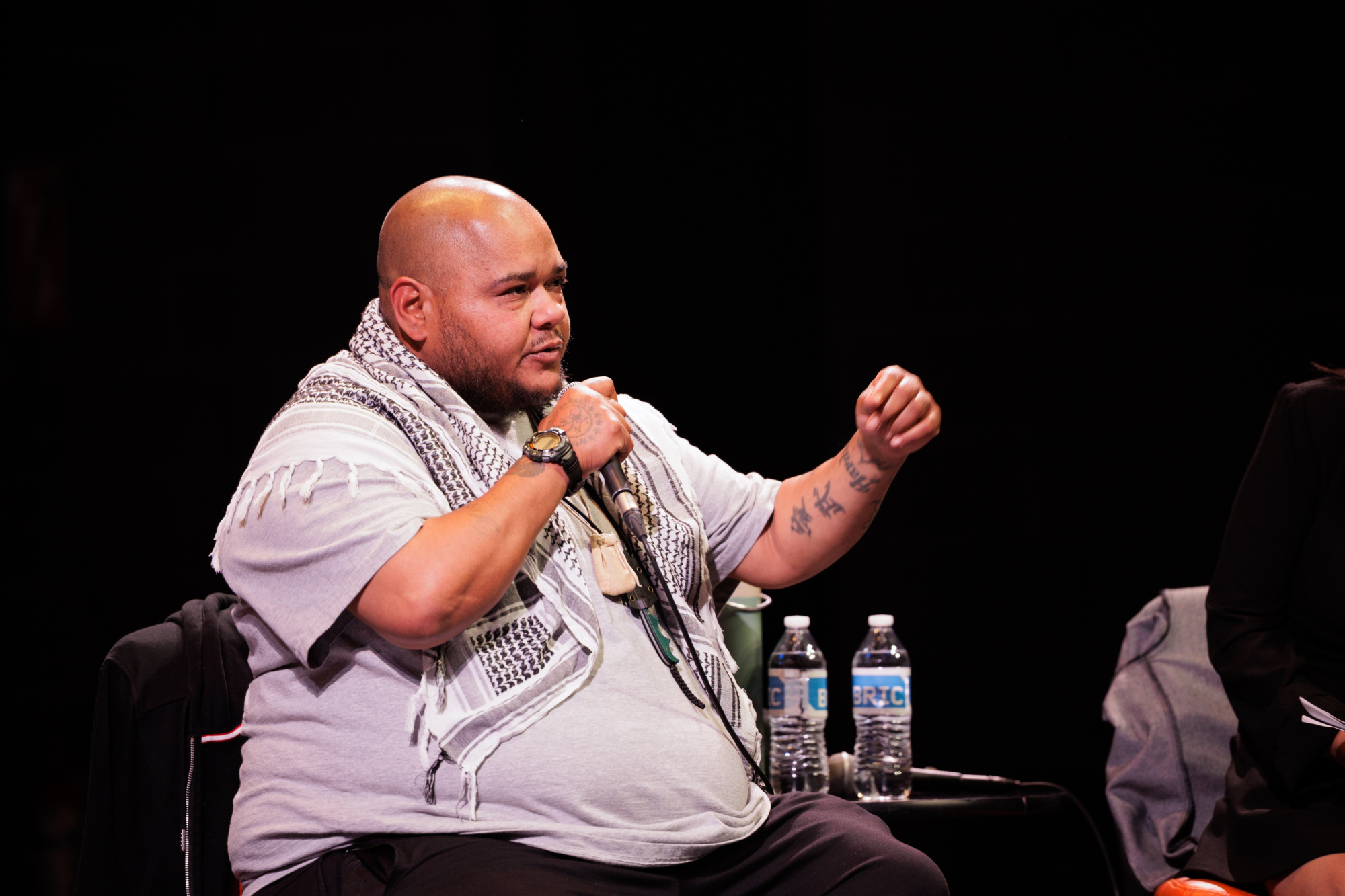 A person with a shaved head and tattoos speaks into a microphone while seated onstage, with two water bottles on the table beside them.