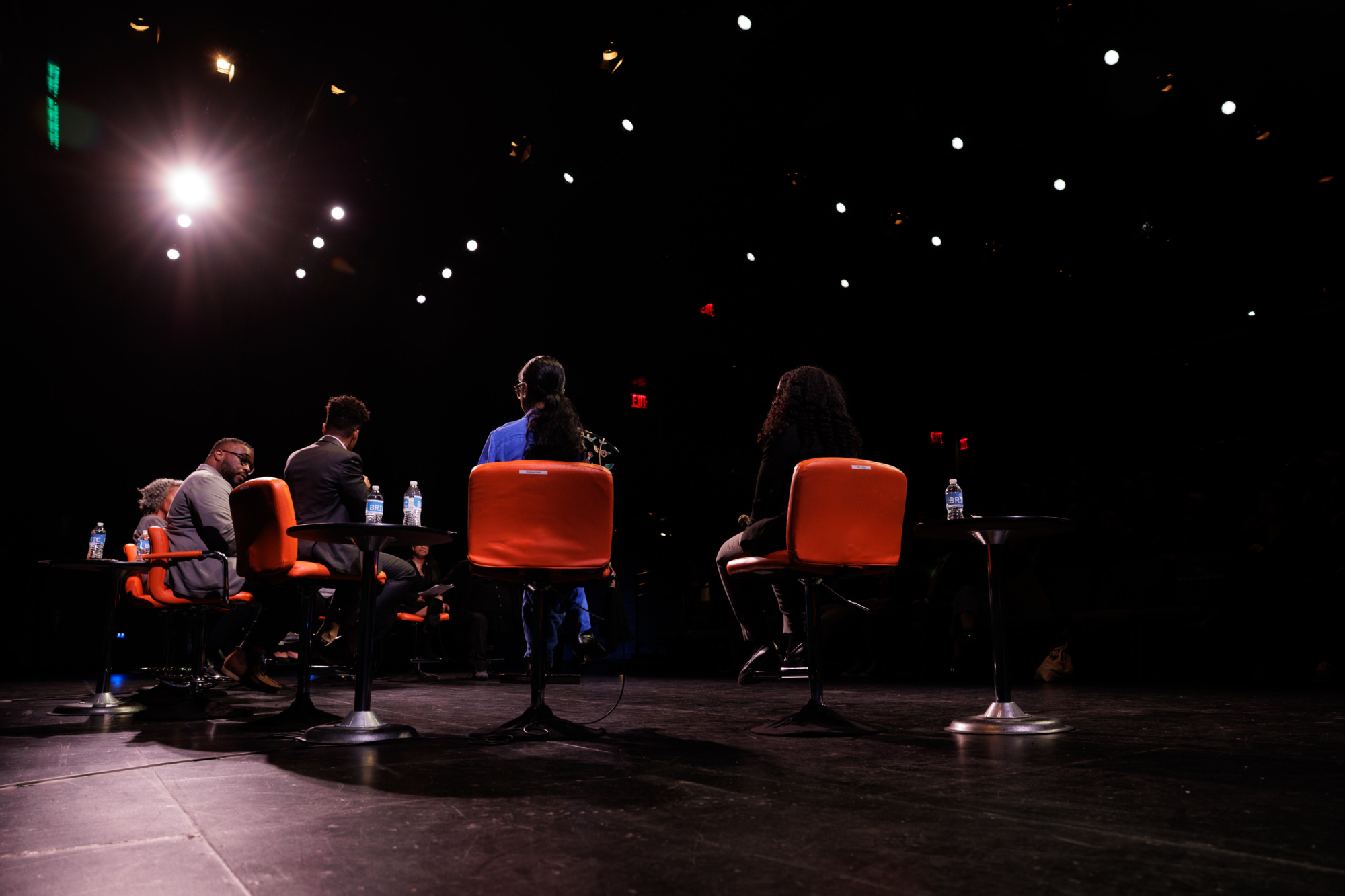 Five people sit on stage in orange chairs facing an audience, illuminated by stage lights. Small tables with bottled water are placed beside each chair.
