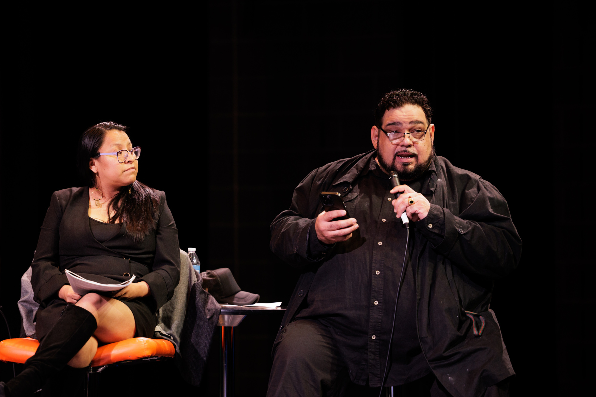 Two people sit on stage; the woman on the left holds papers, while the man on the right speaks into a microphone and looks at his phone.