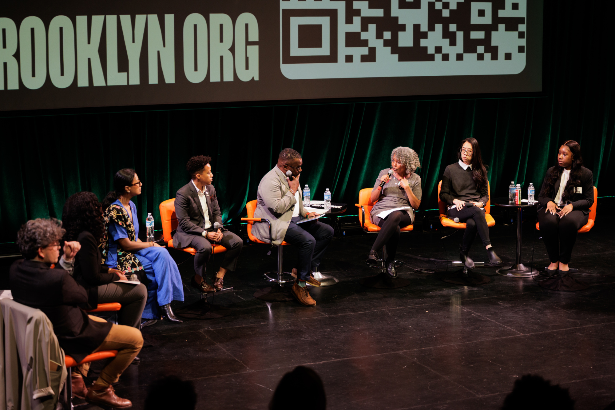 Seven people sit on stage in a panel discussion, each with a microphone, water bottles visible, and a large screen behind them displaying text and a QR code.