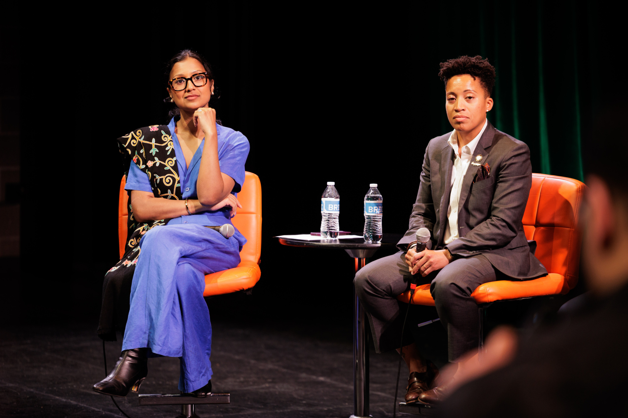 Two people sit on orange chairs on stage with microphones and water bottles, appearing to participate in a discussion or panel event.