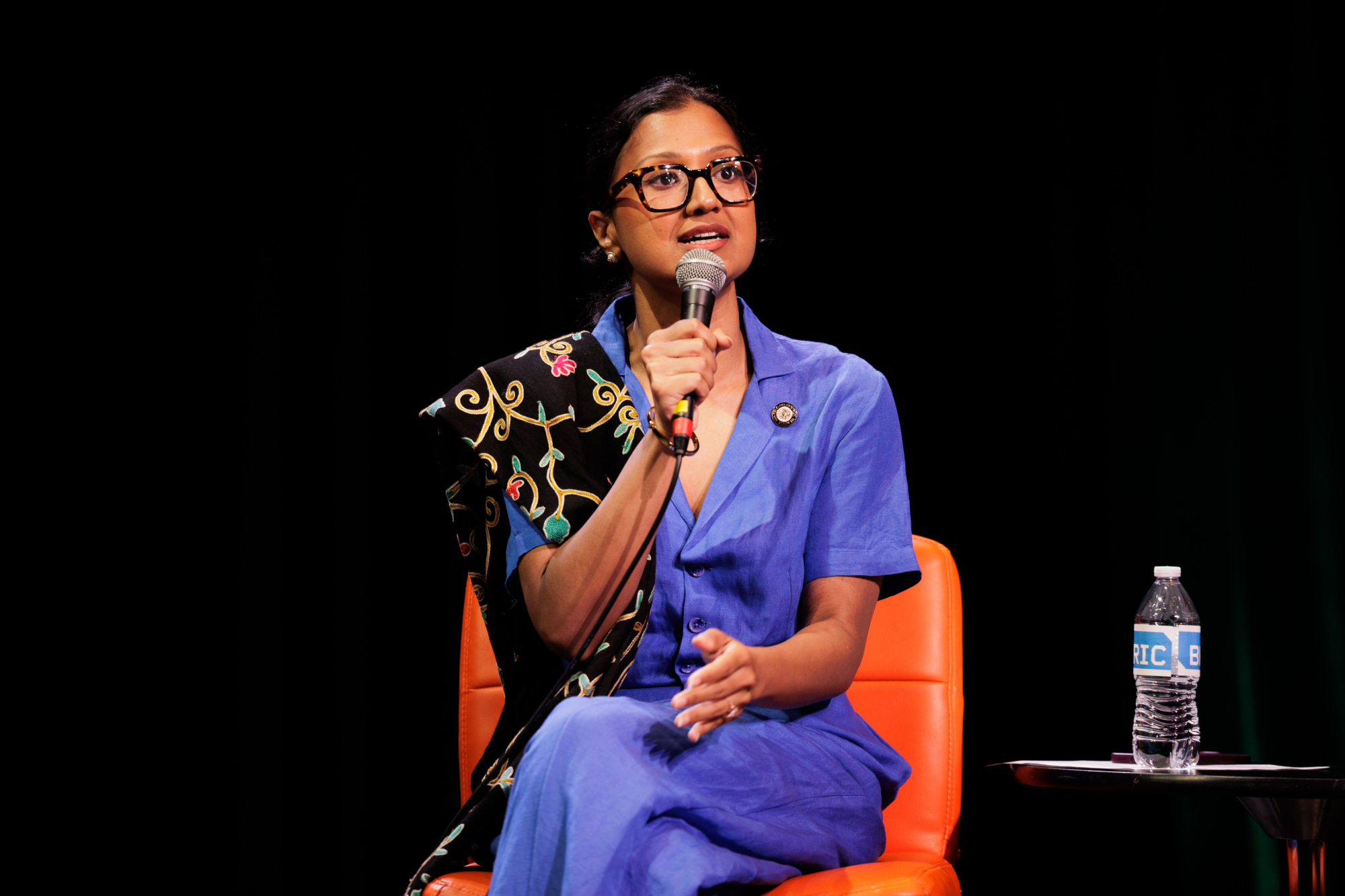 A person in a blue outfit sits on an orange chair, holding a microphone and speaking, with a water bottle on a table nearby against a black background.