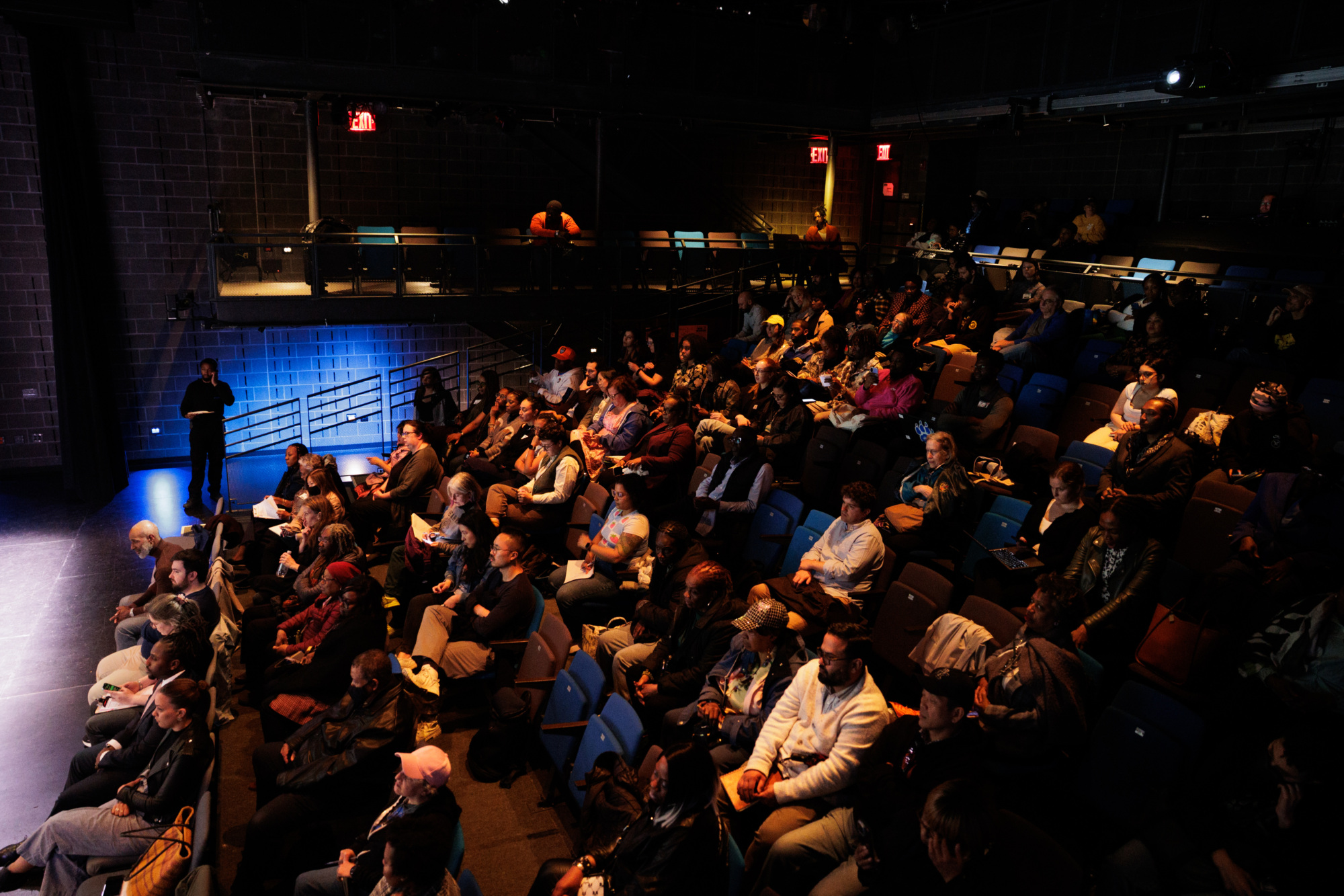 An audience sits in a dimly lit theater, facing a stage on the left, with a person standing near the wall and some empty seats visible among the crowd.